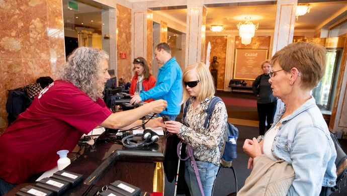 Bezoekers met een visuele beperking krijgen in een hal van een theater audioapparatuur uitgereikt door een medewerker van Komt het Zien. 