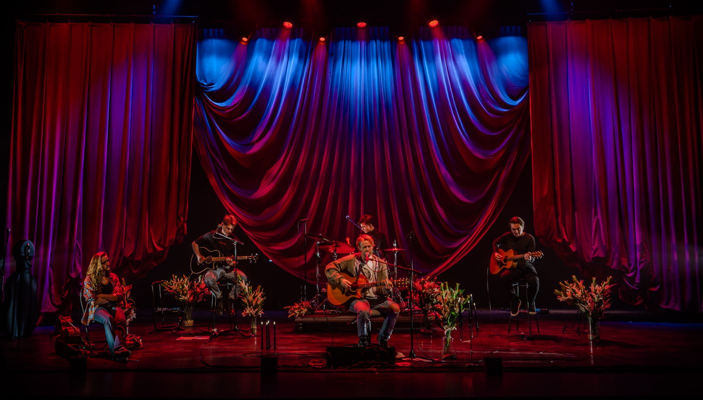 Een band zit op het podium voor een groot gordijn. Tussen hen in staan er planten. Vooraan zit een man met een gitaar voor een microfoon en hij zingt.