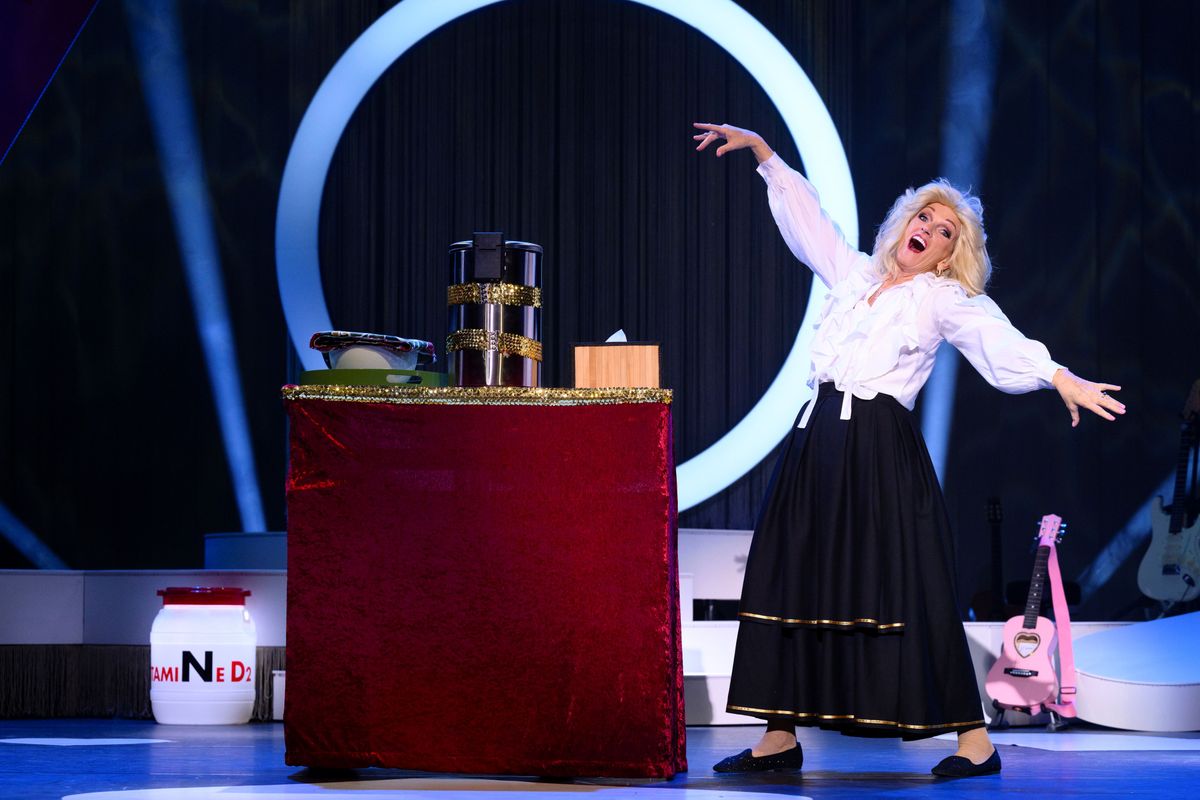 Tineke heeft haar armen wijd en heeft een zwarte rok aan en een witte blouse. Naast haar staat een roze ukelele en een tafeltje met keukenspullen erop. De achtergrond is zwart en er is een witte cirkel te zien.