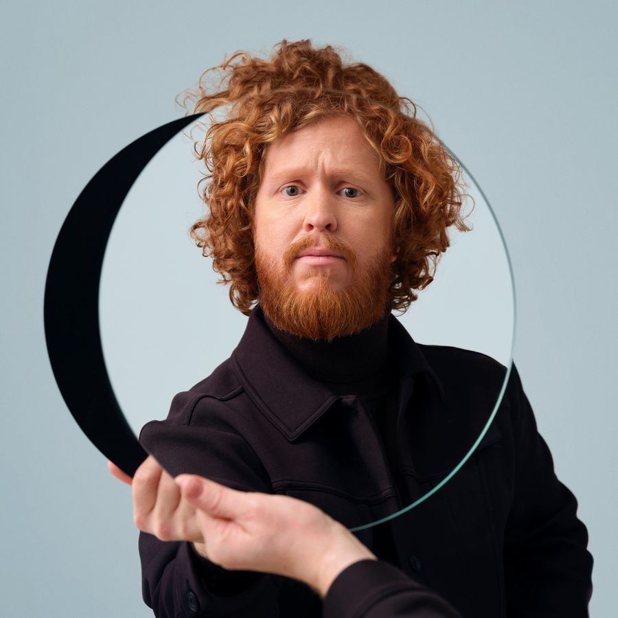 Man in zwart en bos krullen houdt zichzelf een spiegel voor. 