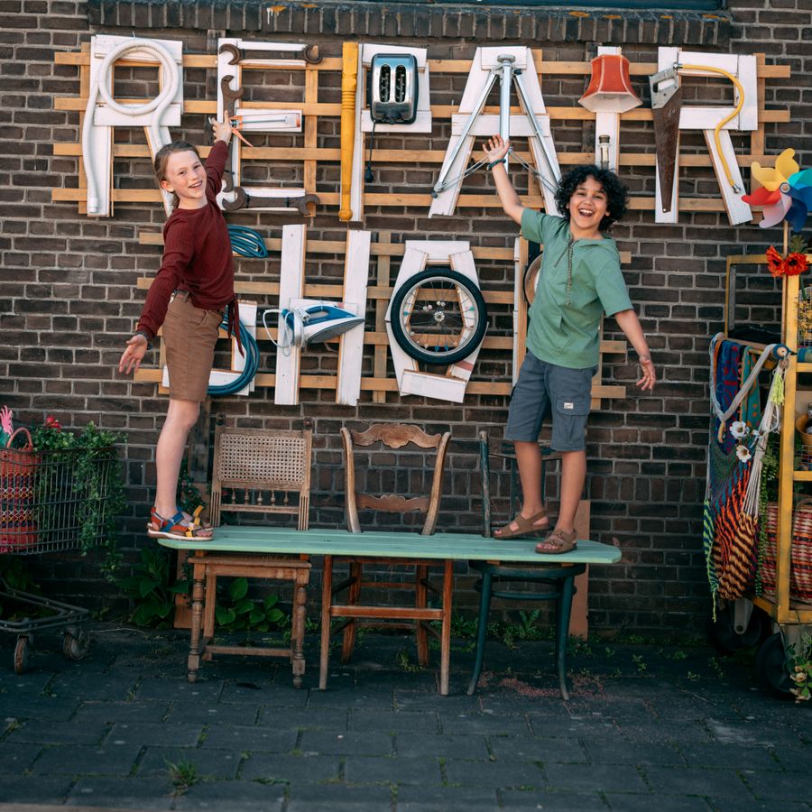 Een jongen en een meisje staan op een plank die op drie stoelen ligt, met hun handen in de lucht. Op de achtergrond is een muur te zien met ‘Repair Shop’, gemaakt van gerecycled materiaal.