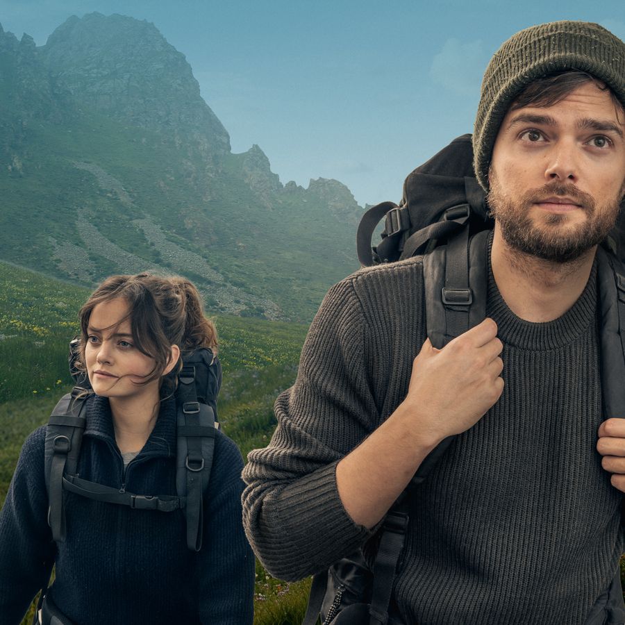 Man met wandelrugzak op de voorgrond kijkend in de camera,  achter hem een een vrouw gekleed in blauw, ook met wandelrugzak, wandelend door een groen bergachtig gebied. 