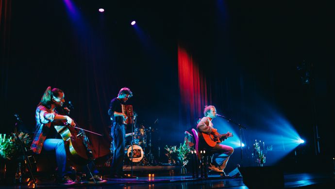 Er staat een band op een podium. Een man met een gitaar zit op een krukje voor een microfoon en zingt. De achtergrond is donker en er hangt een rood theatergordijn.