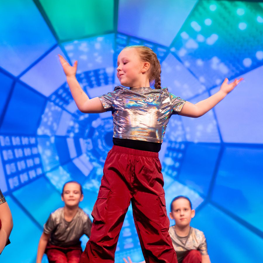 Een meisje van basisschool leeftijd staat te dansen. Andere meisjes zitten daaromheen. Ze heeft een zilveren shirt aan en een rode broek. De achtergrond is blauw.