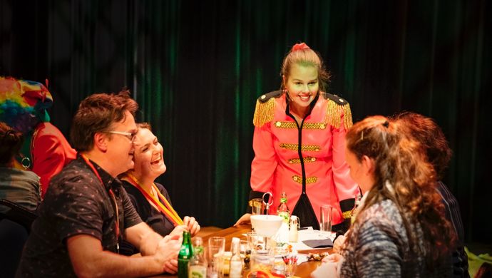Een groepje mensen zitten aan een tafel en lachen. Op tafel liggen allemaal papieren. Naast de tafel staat een vrouw in een roze pak en kijkt met de mensen mee.