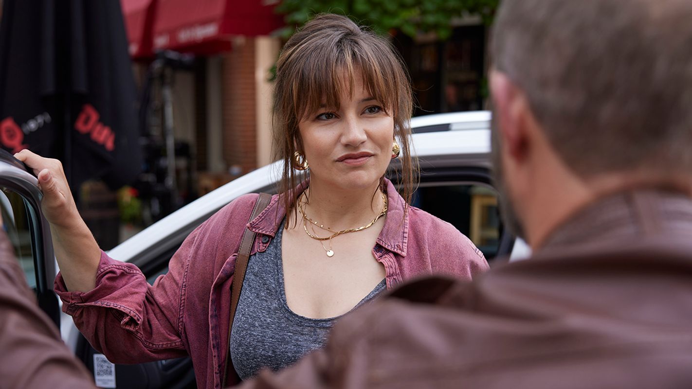 Vrouw met donkere haren, rode blouse en grijs tshirt stapt een auto uit . Haar hand rust op de bovenkant van het portier. 