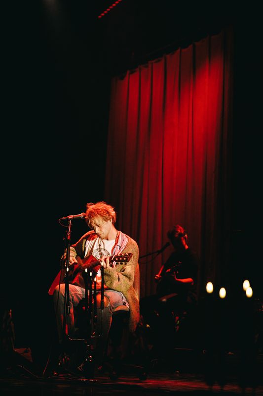 Op de foto zit er een man op het podium voor een rood theatergordijn met zijn gitaar op een krukje voor een microfoon. Hij kijkt naar zijn gitaar.