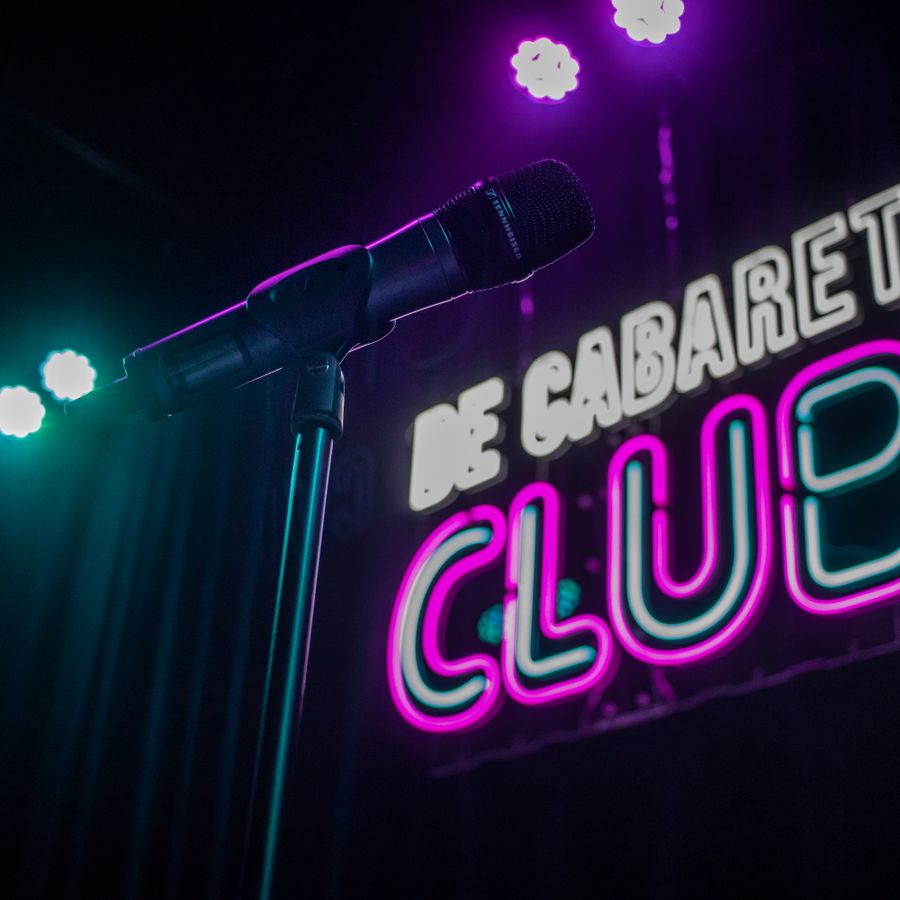 Er is een foto gemaakt van een podium met op de voorgrond een microfoon. Achter het podium staan de woorden: De cabaret club.