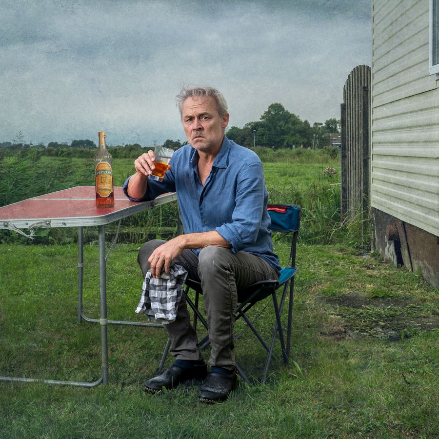 Marcel zit naast een muurtje in een soort weiland aan een campingtafeltje op een campingstoel. Hij houdt een theedoek in zijn handen en drinkt uit een glas. Op tafel naast hem staat een fles drank. Hij kijkt ernstig de camera in.