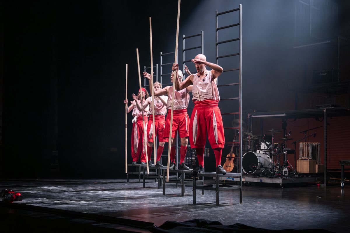 Vier mannen in een rode pak staan ladders op een podium. Achter hen staan er instrumenten.