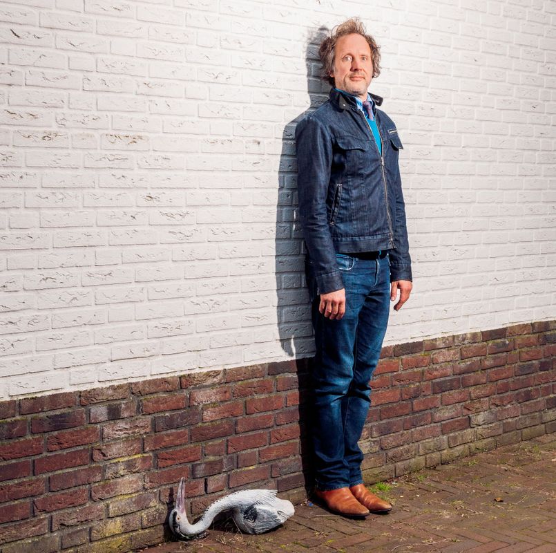 Ronald staat tegen een witte muur. Hij kijkt serieus naar de camera en staat met zijn armen langs zijn lijf, stijf tegen de muur aan. Op de grond ligt er een reiger.