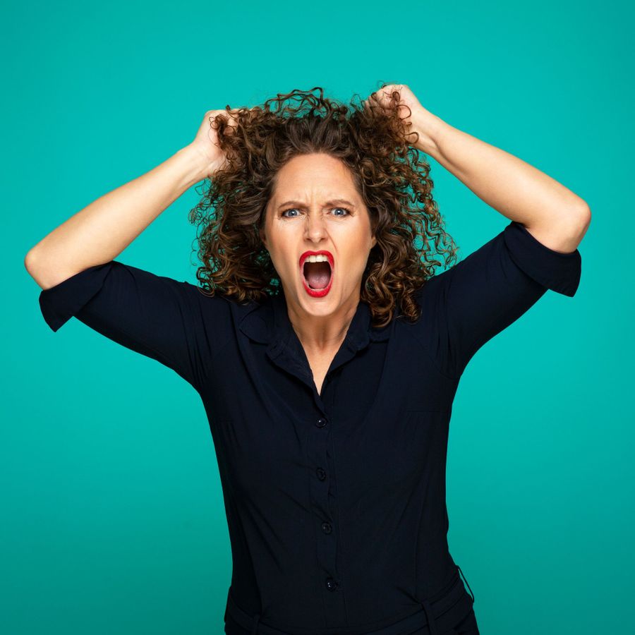 Een vrouw kijkt boos de camera in en heeft haar handen in haar haren. Ze heeft een blauwe blouse aan en rode lippenstift op. Ze staat voor een groene achtergrond