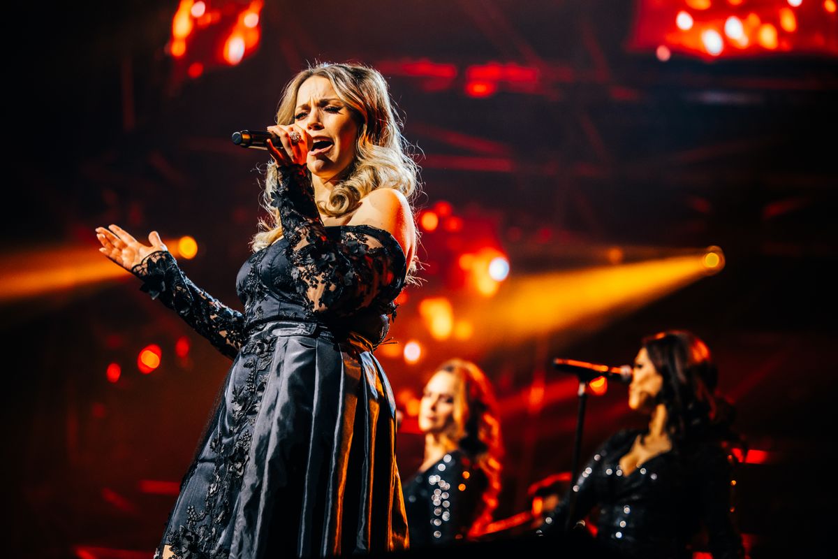 Een vrouw in een zwarte jurk houdt een microfoon vast terwijl ze zingt. Ze heeft haar ogen gesloten. De achtergrond is rood en je ziet achtergrondzangeressen staan.