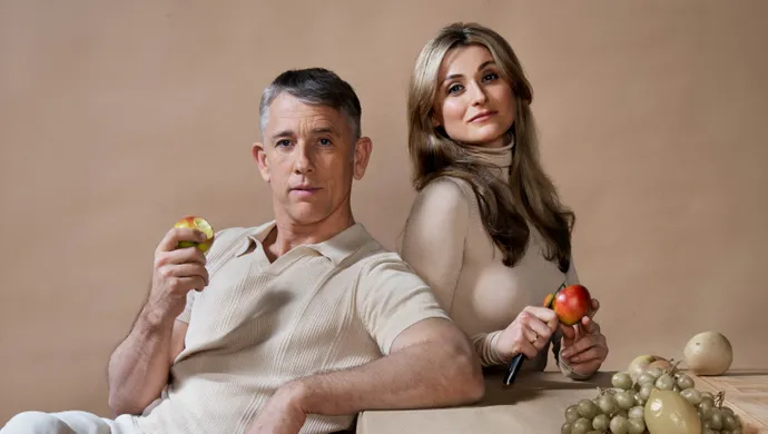 Een man en een vrouw zitten leunend aan een tafel. De man eet een appel en kijkt serieus naar de camera. De vrouw snijdt een appel en kijkt ook serieus naar de camera. Op tafel ligt er groen fruit en de tafel, de achtergrond en hun kleding is beige.