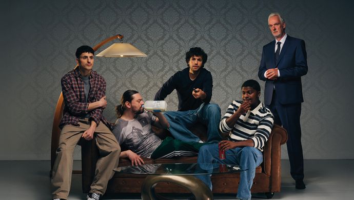 Vier mensen zitten op de bank en kijken wat voor zich uit alsof ze zich vervelen. Naast de bank staat er een man in een blauw pak. Ze staan voor een grijze achtergrond met oud behang.