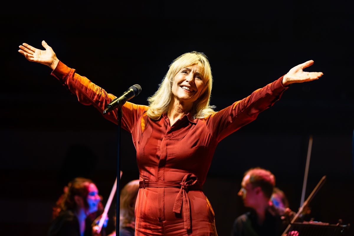 Angela staat op het podium achter een microfoon en lacht. Ze heeft haar handen omhoog.