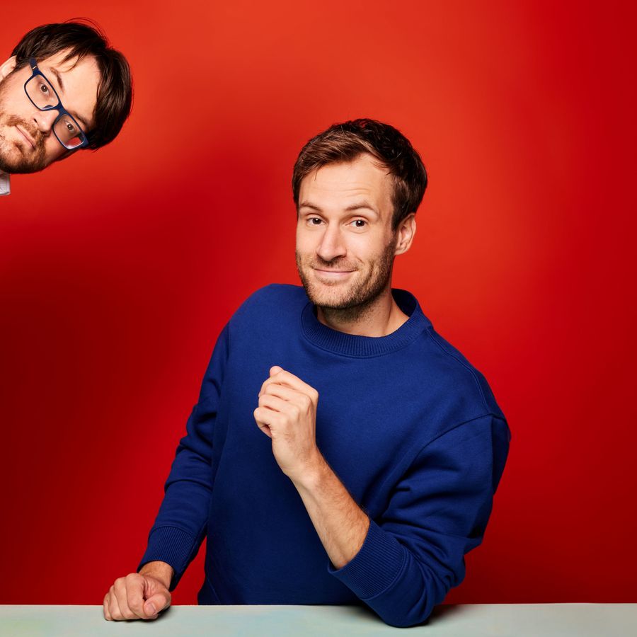 Een man in een blauwe trui kijkt met een glimlach naar de camera. Hij zit voor een rode achtergrond. Aan de zijkant van de afbeelding komt een hoofd van een man met een bril tevoorschijn. Hij heeft een zwart pak aan.