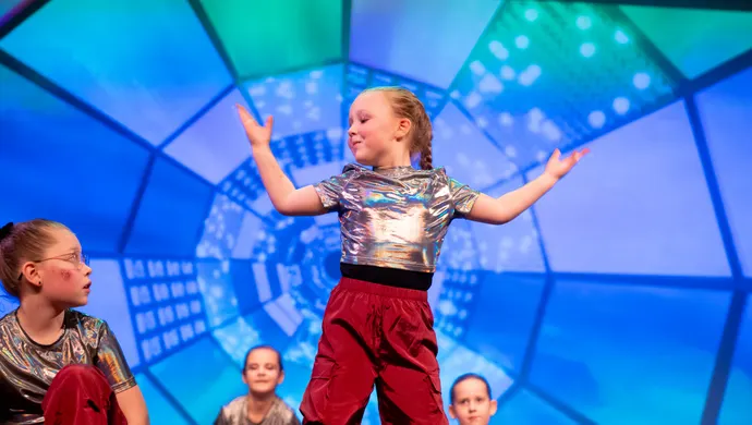 Een meisje van basisschool leeftijd staat te dansen. Andere meisjes zitten daaromheen. Ze heeft een zilveren shirt aan en een rode broek. De achtergrond is blauw.