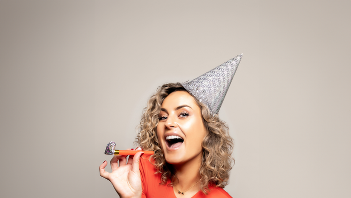 Iris kijkt lachend de camera in en houdt een toeter vast voor haar gezicht. Ze heeft een oranje shirt aan en een hoedje op haar hoofd. De achtergrond is grijs.