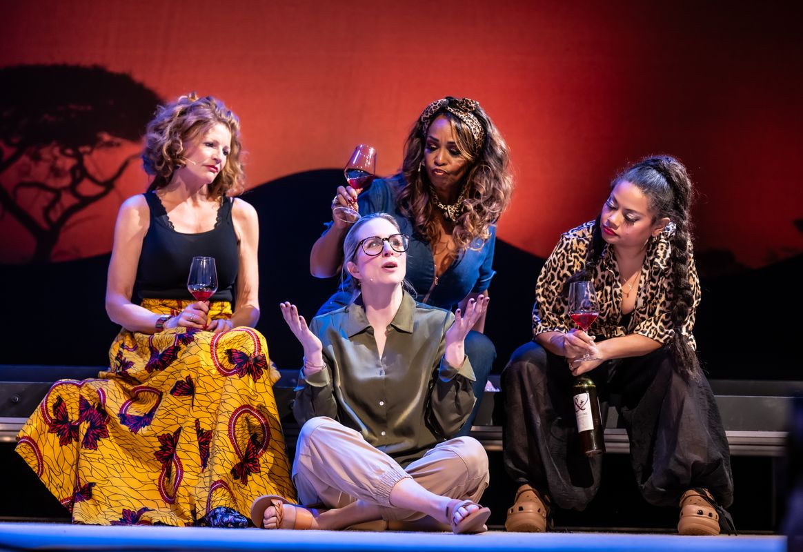 vier dames in fleurige kleding zitten met een wijntje in hun hand. 