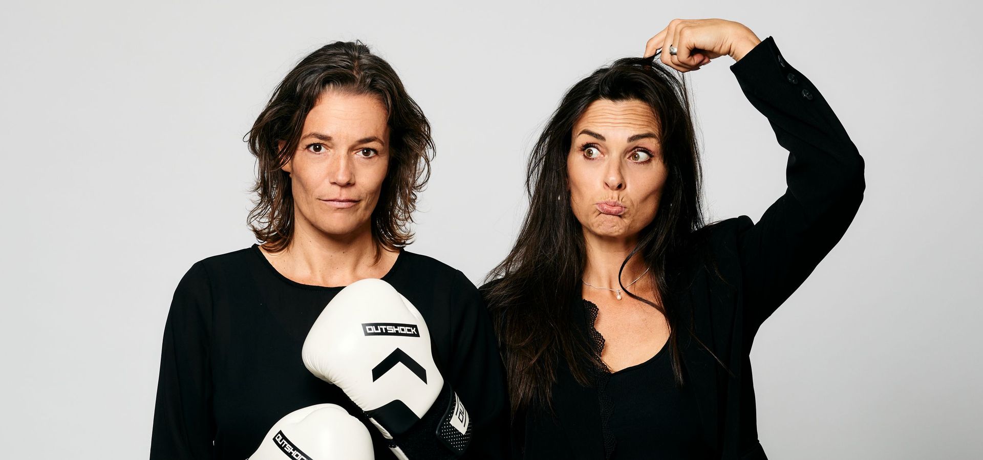 Twee vrouwen staan in zwarte kleding naast elkaar. De vrouw links staat met bokshandschoenen en kijkt serieus de camera in en de vrouw rechts heeft haar vingers op haar hoofd en kijkt verbaasd naar de zijkant.